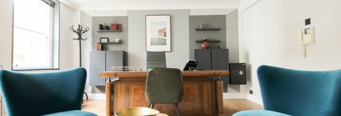 Modern office with a wooden desk, green chair, and two teal armchairs. Perfect for company lets London, the space features shelves and decor on the back wall, sunlight from a large window, and a marble table between the chairs.