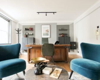 Modern office with a wooden desk, green chair, and two teal armchairs. Perfect for company lets London, the space features shelves and decor on the back wall, sunlight from a large window, and a marble table between the chairs.