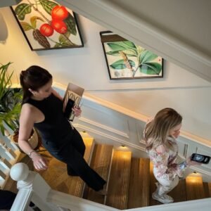 Two women walk up a wooden staircase inside a house, holding books and a drink. The stairway, managed by Elite Property London, features botanical artwork, white panelling, and warm lighting along the steps.