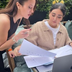 Two women collaborate outdoors, reviewing documents and discussing details about serviced flats. With a laptop, papers, and greenery in the background, they focus on guaranteed rent options for Elite Property London clients.