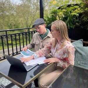 A man and woman sit at an outdoor table with a laptop and documents, discussing Elite Property London company lettings. Green trees and plants surround them, and they appear focused and engaged.