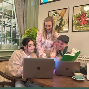Three people sit at a wooden table with two laptops and a notebook, discussing serviced flats. One stands, smiling and pointing at a piece of paper, while the others look engaged. Plants, artwork, and a coffee cup fill the cosy, well-lit background.