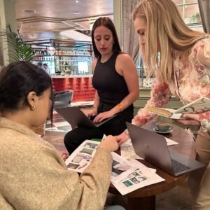Three women collaborate at a table with laptops and design sheets in a stylish, modern café. They’re focused on discussing materials for Elite Property London’s serviced flats, bringing creative ideas to life.