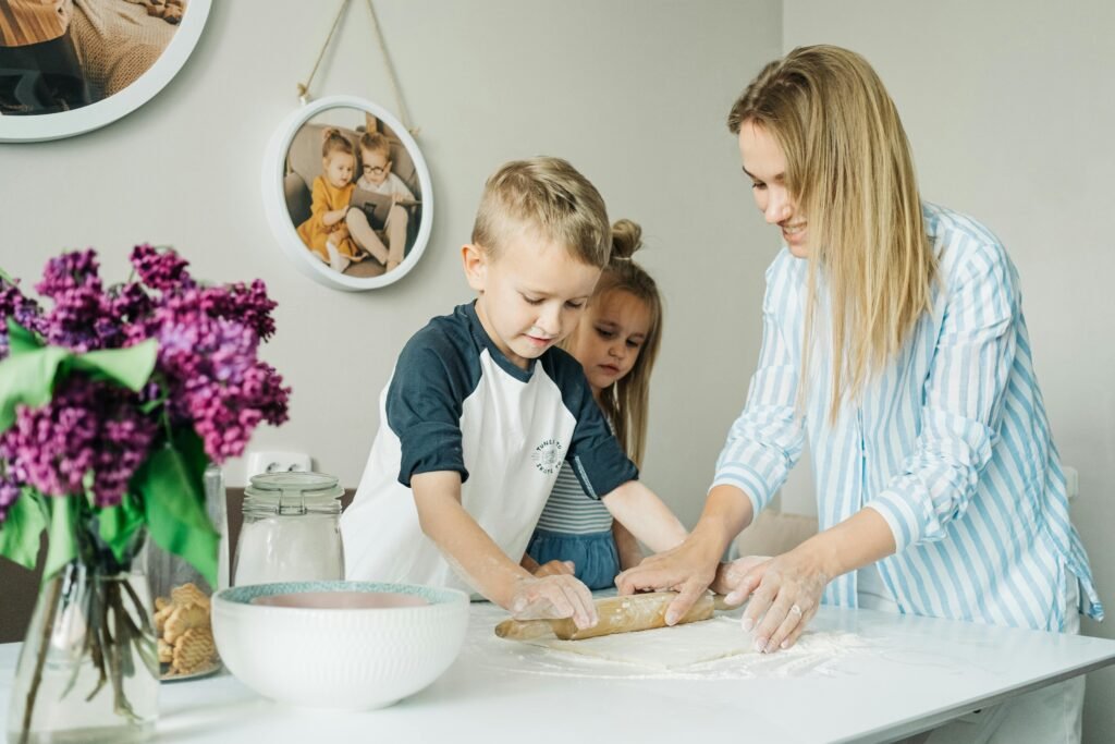 A woman and two children roll out dough together on a white kitchen table in their Elite Property London serviced flat. The table holds a glass jar, a bowl, and purple flowers; family photos hang on the wall in the background.